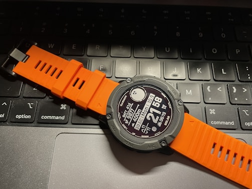 A digital smartwatch with an orange strap is resting on a laptop keyboard. The watch face displays various metrics and information, and the keyboard keys are visible beneath it.
