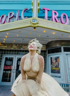 A statue of a woman in a flowing cream dress stands in front of a theater marquee with retro-style letters that spell out 'TROPIC.' The woman is posed with a joyful expression, lips painted bright red and eyes closed. The theater signage above her head features a small clock, and small light bulbs illuminate the marquee. Posters and signs are visible in the windows, with one poster partially reading 'CARLOS.' The colors are vibrant, with accents of turquoise and pink on the marquee.