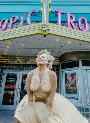 A statue of a woman in a flowing cream dress stands in front of a theater marquee with retro-style letters that spell out 'TROPIC.' The woman is posed with a joyful expression, lips painted bright red and eyes closed. The theater signage above her head features a small clock, and small light bulbs illuminate the marquee. Posters and signs are visible in the windows, with one poster partially reading 'CARLOS.' The colors are vibrant, with accents of turquoise and pink on the marquee.