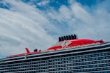 A large cruise ship with a prominent red section on the upper deck and black exhaust funnels. The ship has multiple decks and features the brand name 'Virgin' prominently on the red section. The sky is partly cloudy with patches of blue visible.