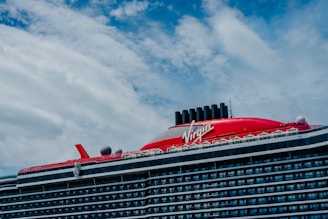 A large cruise ship with a prominent red section on the upper deck and black exhaust funnels. The ship has multiple decks and features the brand name 'Virgin' prominently on the red section. The sky is partly cloudy with patches of blue visible.