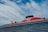 A large cruise ship with a prominent red section on the upper deck and black exhaust funnels. The ship has multiple decks and features the brand name 'Virgin' prominently on the red section. The sky is partly cloudy with patches of blue visible.