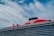 A large cruise ship with a prominent red section on the upper deck and black exhaust funnels. The ship has multiple decks and features the brand name 'Virgin' prominently on the red section. The sky is partly cloudy with patches of blue visible.