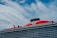 A large cruise ship with a prominent red section on the upper deck and black exhaust funnels. The ship has multiple decks and features the brand name 'Virgin' prominently on the red section. The sky is partly cloudy with patches of blue visible.