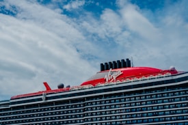 A large cruise ship with a prominent red section on the upper deck and black exhaust funnels. The ship has multiple decks and features the brand name 'Virgin' prominently on the red section. The sky is partly cloudy with patches of blue visible.
