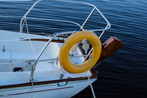 A portion of a sailboat with a yellow life buoy attached to its railing is visible. The boat is near dark blue water, with ropes and metal railings prominent in the scene. The wood trim and clean white surface of the boat provide a striking contrast to the deep color of the water.