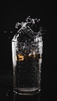 A clear glass of water with ice cubes splashing, captured in motion against a dark background. Water droplets are visibly suspended in mid-air, creating a dynamic and refreshing effect.