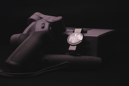 A sleek, ergonomically designed stabilizer or gimbal rests against its packaging, highlighted by soft lighting that emphasizes the matte black and metallic textures. The branding logo is subtly visible on both the device and the box, suggesting a focus on quality and technology.