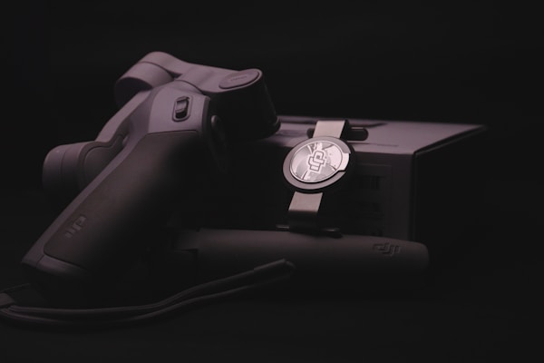 A sleek, ergonomically designed stabilizer or gimbal rests against its packaging, highlighted by soft lighting that emphasizes the matte black and metallic textures. The branding logo is subtly visible on both the device and the box, suggesting a focus on quality and technology.