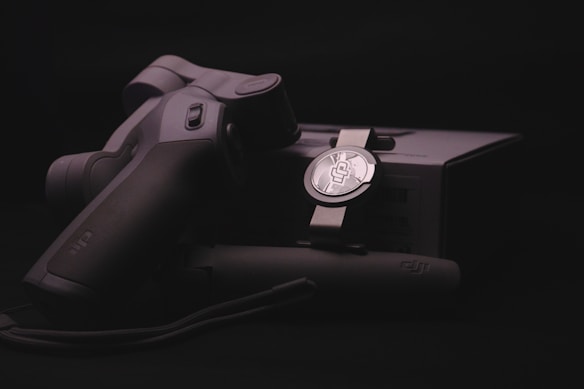 A sleek, ergonomically designed stabilizer or gimbal rests against its packaging, highlighted by soft lighting that emphasizes the matte black and metallic textures. The branding logo is subtly visible on both the device and the box, suggesting a focus on quality and technology.