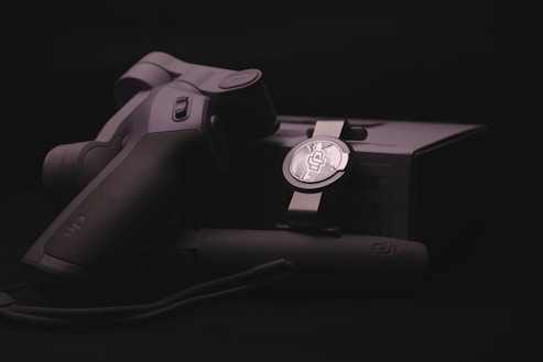 A sleek, ergonomically designed stabilizer or gimbal rests against its packaging, highlighted by soft lighting that emphasizes the matte black and metallic textures. The branding logo is subtly visible on both the device and the box, suggesting a focus on quality and technology.
