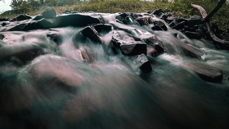 A flowing stream moves over a collection of dark, smooth stones. The motion of the water creates a blurred effect, highlighting the movement of the current. Surrounding the stream are patches of green grass and some trees in the background.