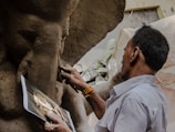 A person is sculpting a statue from clay, referencing an image held in their hand. The photo captures the intricate details of the sculpting process, particularly the hands as they work. The setting suggests a workshop with natural light illuminating the scene.