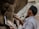 A person is sculpting a statue from clay, referencing an image held in their hand. The photo captures the intricate details of the sculpting process, particularly the hands as they work. The setting suggests a workshop with natural light illuminating the scene.