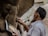 A person is sculpting a statue from clay, referencing an image held in their hand. The photo captures the intricate details of the sculpting process, particularly the hands as they work. The setting suggests a workshop with natural light illuminating the scene.