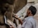 A person is sculpting a statue from clay, referencing an image held in their hand. The photo captures the intricate details of the sculpting process, particularly the hands as they work. The setting suggests a workshop with natural light illuminating the scene.
