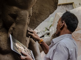 A person is sculpting a statue from clay, referencing an image held in their hand. The photo captures the intricate details of the sculpting process, particularly the hands as they work. The setting suggests a workshop with natural light illuminating the scene.