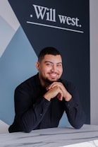 A smiling person with short hair and a beard leans on a marble-patterned surface. They are wearing a black dress shirt. Behind them is a dark blue and white geometric wall design with the text 'Will west.'