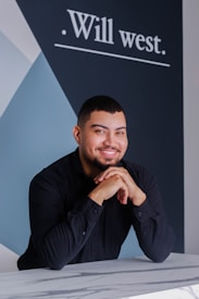 A smiling person with short hair and a beard leans on a marble-patterned surface. They are wearing a black dress shirt. Behind them is a dark blue and white geometric wall design with the text 'Will west.'