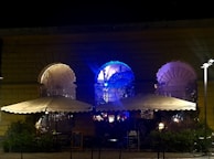 A nighttime scene depicting an outdoor seating area of a café or restaurant. The venue is built with large arched windows, each illuminated with colorful lights, casting a glow in shades of blue and purple. Two large umbrellas provide cover over the seating area, and small tables and chairs are arranged beneath them. A chalkboard sign is visible near the entrance, suggesting live music or events. The mood is set with ambient lighting, creating a cozy and inviting atmosphere.