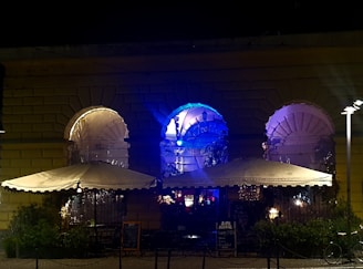 A nighttime scene depicting an outdoor seating area of a café or restaurant. The venue is built with large arched windows, each illuminated with colorful lights, casting a glow in shades of blue and purple. Two large umbrellas provide cover over the seating area, and small tables and chairs are arranged beneath them. A chalkboard sign is visible near the entrance, suggesting live music or events. The mood is set with ambient lighting, creating a cozy and inviting atmosphere.