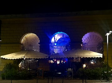 A nighttime scene depicting an outdoor seating area of a caf&eacute; or restaurant. The venue is built with large arched windows, each illuminated with colorful lights, casting a glow in shades of blue and purple. Two large umbrellas provide cover over the seating area, and small tables and chairs are arranged beneath them. A chalkboard sign is visible near the entrance, suggesting live music or events. The mood is set with ambient lighting, creating a cozy and inviting atmosphere.