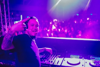 A DJ stands behind a mixing console, wearing headphones and smiling, with colorful lights illuminating the background. The crowd is blurred, contributing to an energetic atmosphere. The DJ gestures with a sign of enthusiasm.