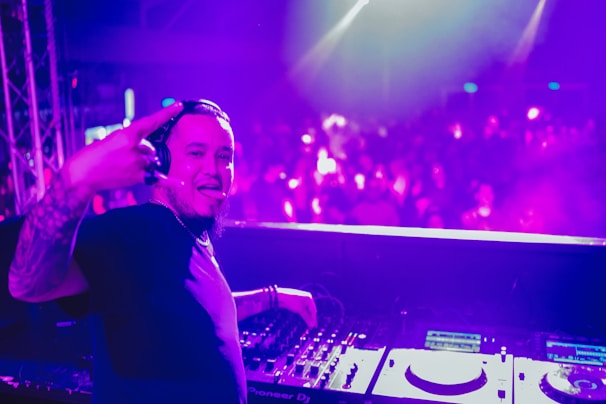 A DJ stands behind a mixing console, wearing headphones and smiling, with colorful lights illuminating the background. The crowd is blurred, contributing to an energetic atmosphere. The DJ gestures with a sign of enthusiasm.