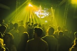 A large crowd of people is gathered in a dimly lit venue, illuminated by yellow and green stage lights. A DJ booth is visible at the front with a large screen displaying visuals including the word 'BASSICK'. The atmosphere suggests a music concert or nightclub event.