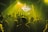 A large crowd of people is gathered in a dimly lit venue, illuminated by yellow and green stage lights. A DJ booth is visible at the front with a large screen displaying visuals including the word 'BASSICK'. The atmosphere suggests a music concert or nightclub event.