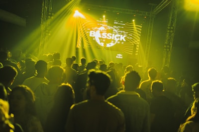 A large crowd of people is gathered in a dimly lit venue, illuminated by yellow and green stage lights. A DJ booth is visible at the front with a large screen displaying visuals including the word 'BASSICK'. The atmosphere suggests a music concert or nightclub event.