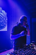 A DJ is performing in a dimly lit club or venue, standing behind a DJ console filled with various controls and buttons. The lighting is predominantly blue and purple, creating a vibrant atmosphere. The DJ is wearing headphones and appears focused on the music.