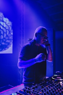 A DJ is performing in a dimly lit club or venue, standing behind a DJ console filled with various controls and buttons. The lighting is predominantly blue and purple, creating a vibrant atmosphere. The DJ is wearing headphones and appears focused on the music.