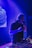 A DJ is performing in a dimly lit club or venue, standing behind a DJ console filled with various controls and buttons. The lighting is predominantly blue and purple, creating a vibrant atmosphere. The DJ is wearing headphones and appears focused on the music.