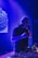 A DJ is performing in a dimly lit club or venue, standing behind a DJ console filled with various controls and buttons. The lighting is predominantly blue and purple, creating a vibrant atmosphere. The DJ is wearing headphones and appears focused on the music.