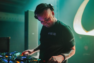 A DJ wearing headphones is deeply engaged with a set of DJ equipment, surrounded by dark ambient lighting and subtle stage lights. The person is smiling and focused, creating a lively and energetic atmosphere.