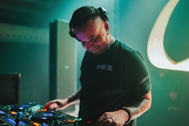 A DJ wearing headphones is deeply engaged with a set of DJ equipment, surrounded by dark ambient lighting and subtle stage lights. The person is smiling and focused, creating a lively and energetic atmosphere.