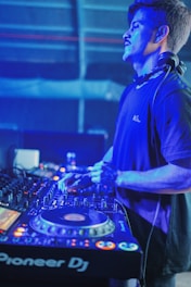 A DJ wearing headphones is focused on mixing tracks at a DJ console in a dimly lit environment with blue and purple lighting. The professional DJ equipment is prominently featured, showing various knobs, buttons, and a digital display.