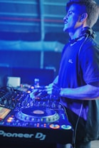 A DJ wearing headphones is focused on mixing tracks at a DJ console in a dimly lit environment with blue and purple lighting. The professional DJ equipment is prominently featured, showing various knobs, buttons, and a digital display.