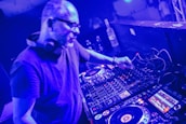 A person is using DJ equipment, adjusting controls on a mixing console in a dimly lit setting. Headphones are around their neck, and several buttons and displays on the equipment are illuminated.