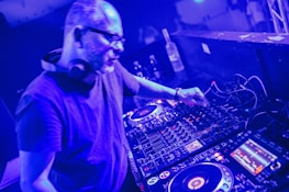 A person is using DJ equipment, adjusting controls on a mixing console in a dimly lit setting. Headphones are around their neck, and several buttons and displays on the equipment are illuminated.