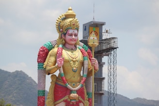A large, colorful statue of a Hindu deity standing outdoors with a tall tower structure in the background. The statue is adorned with vibrant garlands, intricate jewelry, and a crown. The sky is slightly cloudy and there's a mountain range visible in the distance.