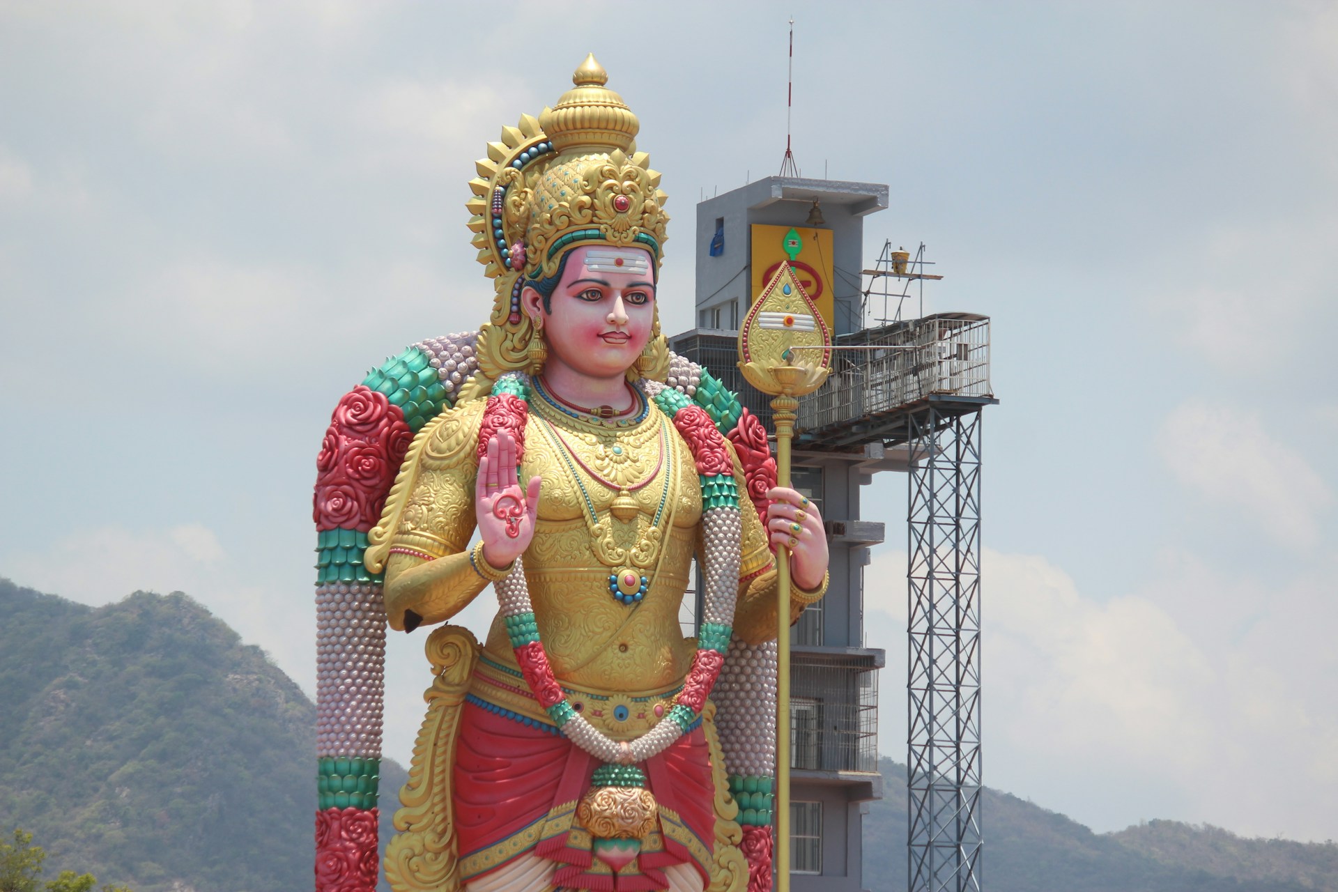 A large, colorful statue of a Hindu deity standing outdoors with a tall tower structure in the background. The statue is adorned with vibrant garlands, intricate jewelry, and a crown. The sky is slightly cloudy and there's a mountain range visible in the distance.