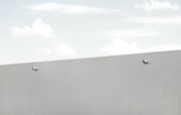A minimalist scene featuring a large, light grey wall with two security cameras attached. The top third of the image shows a pale blue sky with soft, scattered clouds.