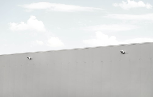 A minimalist scene featuring a large, light grey wall with two security cameras attached. The top third of the image shows a pale blue sky with soft, scattered clouds.