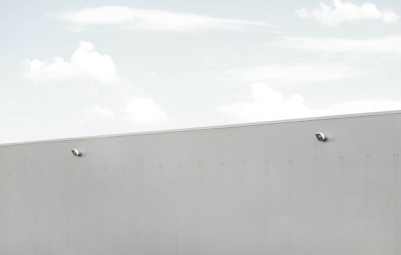 A minimalist scene featuring a large, light grey wall with two security cameras attached. The top third of the image shows a pale blue sky with soft, scattered clouds.