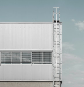 A modern industrial building facade featuring a vertical metal ladder on the right side and horizontal windows with closed blinds. The building exterior is made of light grey metal panels.