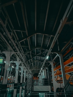 A spacious industrial interior with a high ceiling supported by large metal beams. The lighting is dim, with fluorescent lights casting a greenish hue. The floor appears to be reflective, contributing to a somewhat futuristic atmosphere. Structural elements and equipment line the sides of the space.