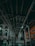 A spacious industrial interior with a high ceiling supported by large metal beams. The lighting is dim, with fluorescent lights casting a greenish hue. The floor appears to be reflective, contributing to a somewhat futuristic atmosphere. Structural elements and equipment line the sides of the space.