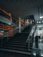 A modern stairway with black steps and metal railings in an indoor setting. The stairway leads to an upper level with a sign above indicating directions and symbols for an elevator and restroom. The surrounding area has an industrial design with orange and silver structural elements and a ceiling with horizontal beams.